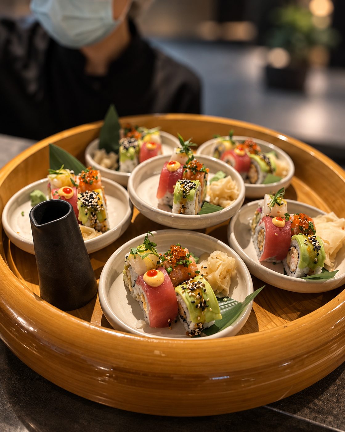 Round wooden tray with small plates of colourful sushi rolls and pickled ginger