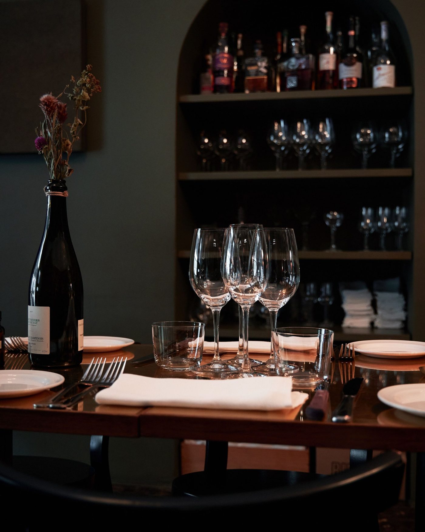 Laid table with wine glasses, folded napkins and a dried flower in a bottle, shelves of glasses behind